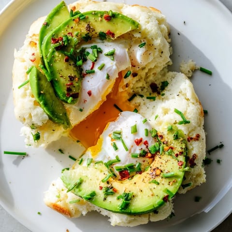 Four golden Cloud Bread Breakfast Clouds sit on a baking sheet, each filled with a baked egg yolk and topped with creamy avocado slices and a poached egg.