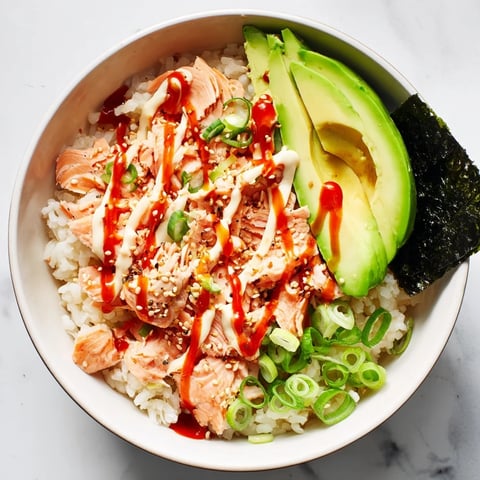 A close-up of a vibrant Emily Mariko Salmon Rice Bowl featuring flaked salmon, creamy avocado slices, and spicy mayo drizzle.