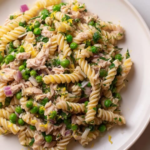 Bright Lemony Tuna Pasta Salad in a white bowl, featuring flaked tuna, green peas, and fresh parsley, served with a lemon wedge.